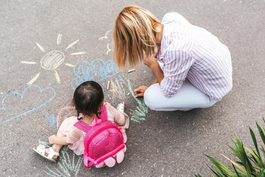Eine junge Frau sitz mit einem kleinen Mädchen, das einen Kindergartenrucksack auf dem Rücken trägt, auf dem Hof und malt mit Kreide auf den Boden
