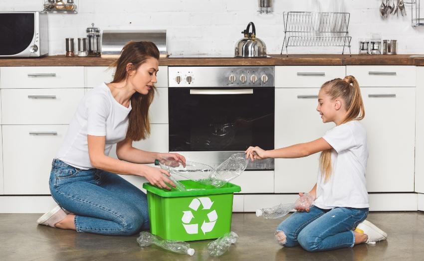 Eine Mutter und ihre Tochter füllen eine grüne box mit Recycling-Logo in ihrer Küche mit leeren Pfandflaschen