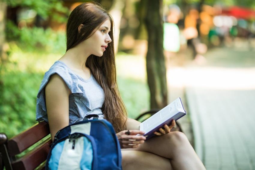 Studentin sitzt auf einer Parkbank und liest, im Vordergrund ein Schulrucksack für Damen