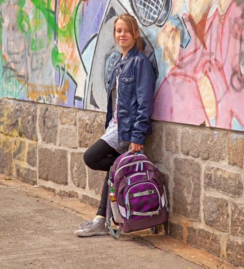 Eine Schülerin lehnt an der Wand der Schule. In der Hand trägt sie ihren Schulrucksack Schultasche für die weiterführende Schule